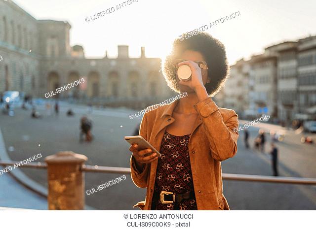 Young woman with afro hair having hot drink, using smartphone in city, Florence, Toscana, Italy