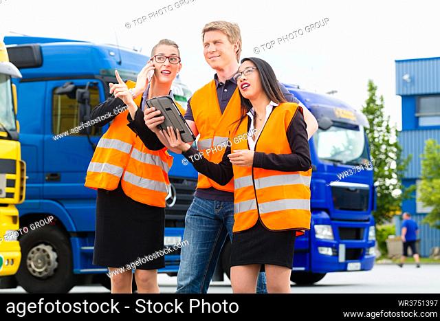 Logistics - proud driver or forwarder and colleagues with tablet computer, in front of trucks and trailers, on a transshipment point
