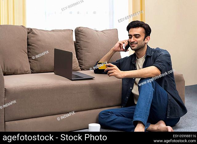 A young man with laptop holding credit card talking on mobile in his living room