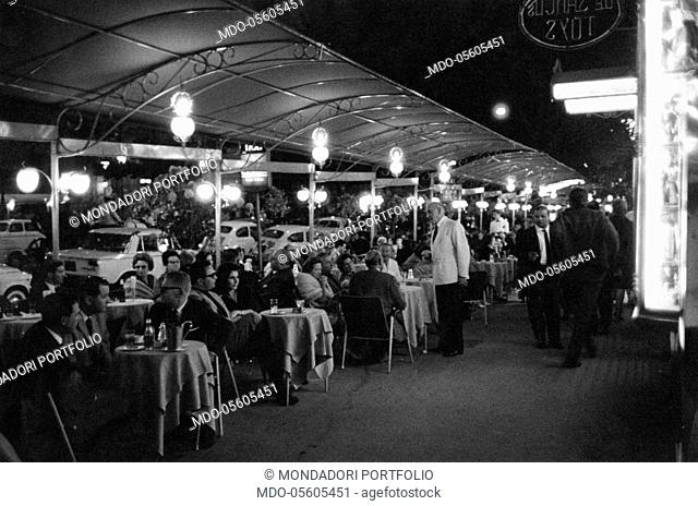 Many people crowding the cafés on via Veneto in the night. Rome, 1963