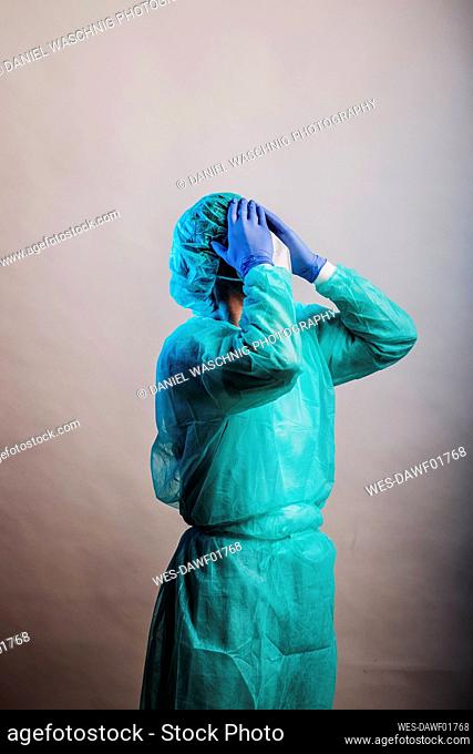 Male doctor wearing protective suit standing with head in hands against gray background