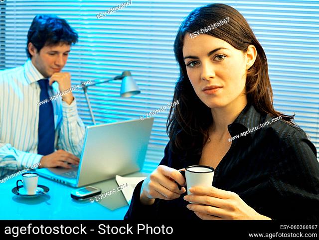 Tired businesswoman drinking coffee during a meeting, businessman working with laptop computer in the background