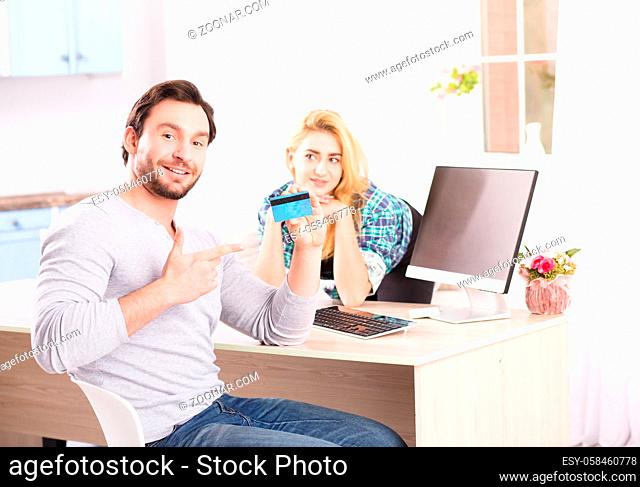 Toned of cheerful young bearded man smiling and showing credit card to camera while sitting in front of computer. Happy blond lady on background