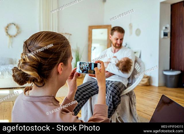 Woman photographing man carrying daughter on chair at home