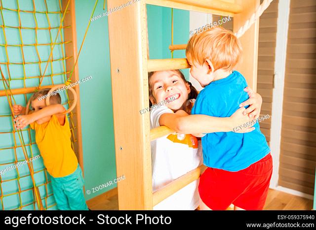 Close view of brothers and sister playing in a wooden home sports complex. Girl widely smiling, holding her brother, helping him to climb wooden bars