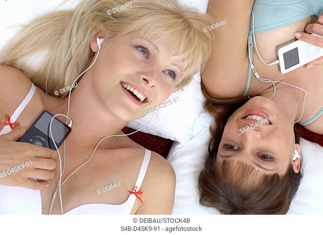 Blond woman lying next to a brunette young woman, both in underwear and listening to music, high angle view, close-up