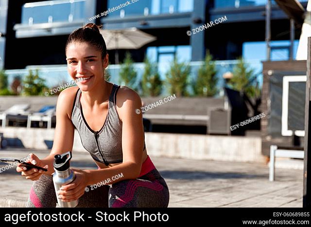 Outdoor shot of attractive fitness woman sitting with smartphone and drinking water