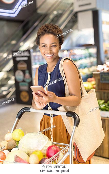 Portrait smiling young woman using cell phone, grocery shopping in market