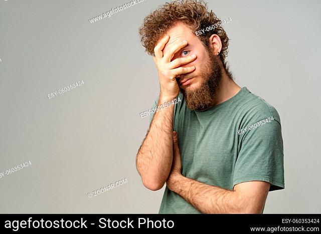 Hiding handsome young man covered his face with hand pry with his eye, curly hair in olive t-shirt isolated on white background