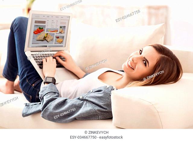 Get some rest. Cheerful delighted beautiful girl lying on the sofa and smiling while using laptop