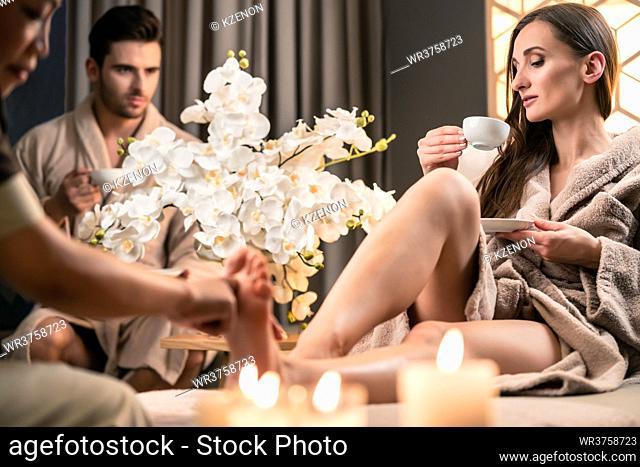 Beautiful woman drinking tea during therapeutic foot massage next to her partner at Asian wellness center
