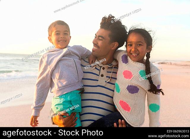 Happy biracial father carrying children while standing at beach against sky during sunset