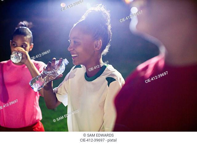 Smiling young female soccer players resting, drinking from water bottles on field at night