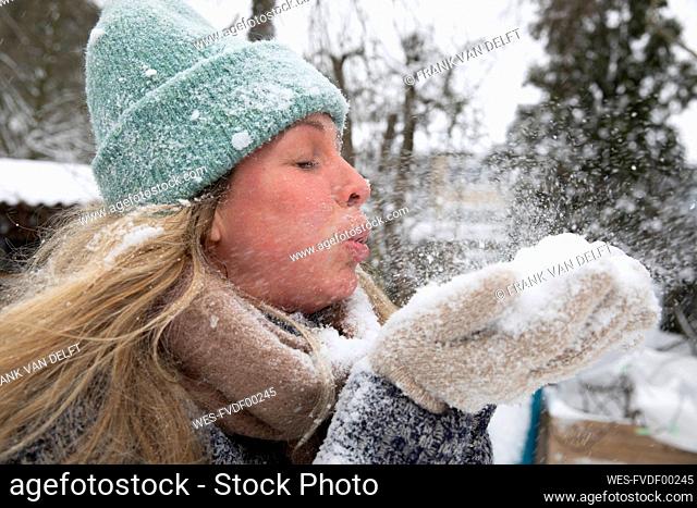 Blond woman with eyes closed blowing snow while standing at backyard during winter