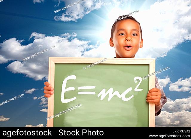 Proud hispanic boy holding chalkboard with theory of relativity over blue sky and sun burst clouds