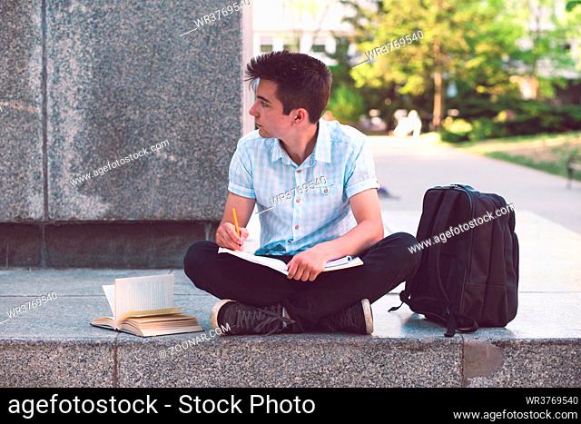 Student making the notes learning from books sitting on a monument outside of university. Young boy wearing a blue shirt and dark jeans