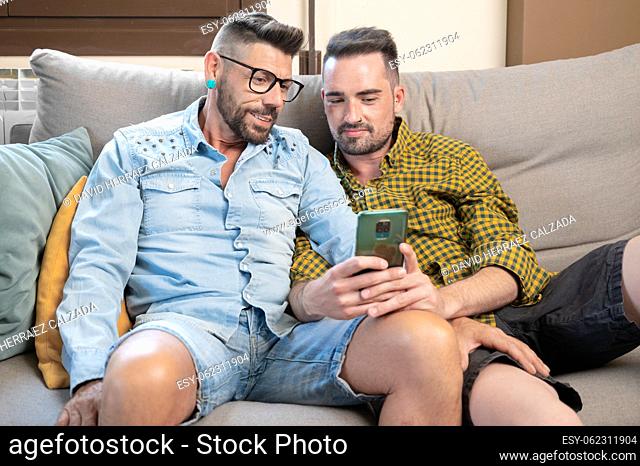 Happy young gay couple using mobile phone while sitting on a sofa in the living room. High quality photography
