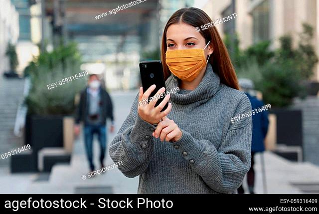 Portrait of young woman with protective mask video calling with someone while walking in city street