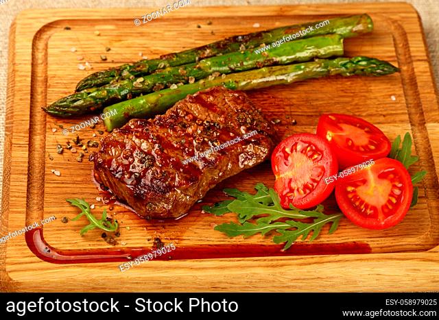 Close up one grilled roasted beef steak with green asparagus and cherry tomatoes on wooden cutting board, high angle view