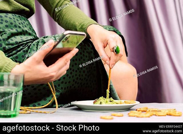 Close-up of woman using smart phone while eating guacamole and pretzel on table at home