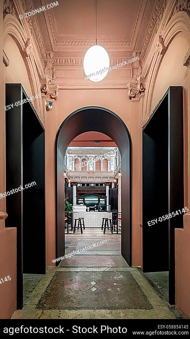Arch black doorway to the hall in a restaurant with walls with stucco molding. There is a tiled rack , wooden sideboard, tables with stools, textured floor