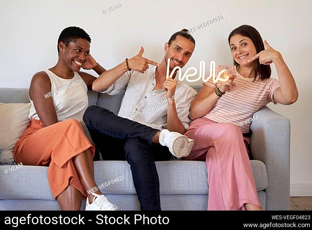 Happy male and female coworkers pointing at Hello sign illuminated text while sitting on sofa