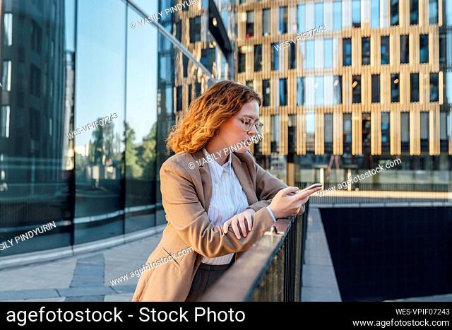 Young redhead businesswoman using mobile phone leaning on railing