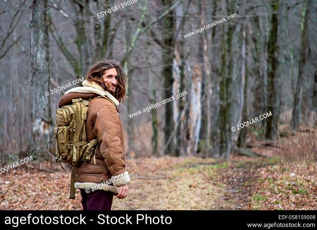 Rear view of a young man with long hair, sheepskin winter coat and green military backpack looking at camera while walking in the woodlands