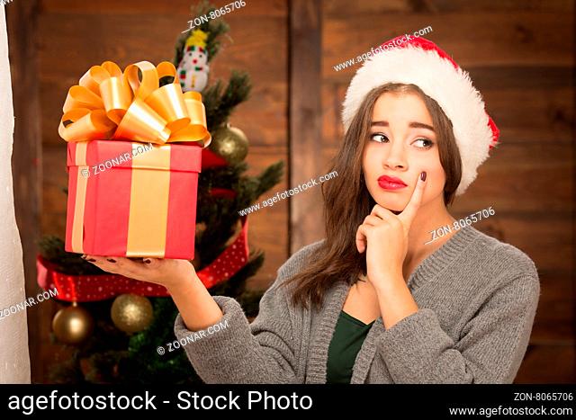 Serious girl holding a present in front of New Year tree and thinking about possible gifts in it. Pretty lady looking away and touching her cheek