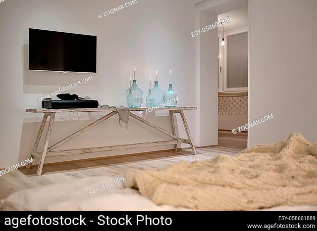 Modern interior with white walls and a parquet. There is a wooden table with candles in bottles, soundbar, shawl, TV, bed with a plaid
