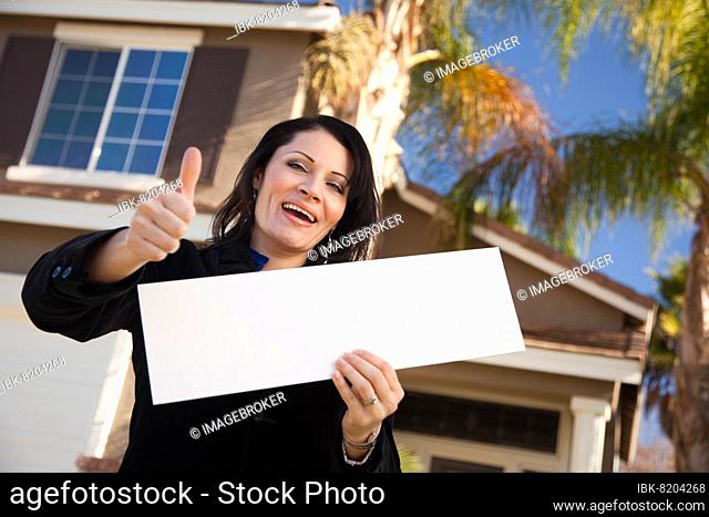 Happy attractive hispanic woman with thumbs up holding blank sign in front of house