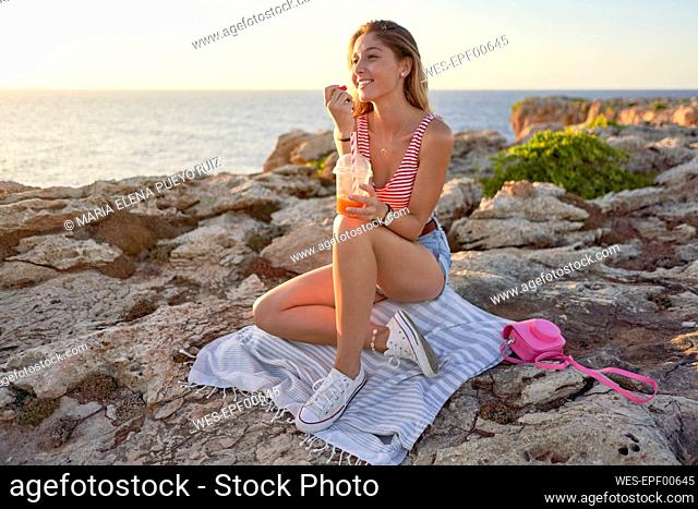 Young woman sitting on rocky beach, eating candies