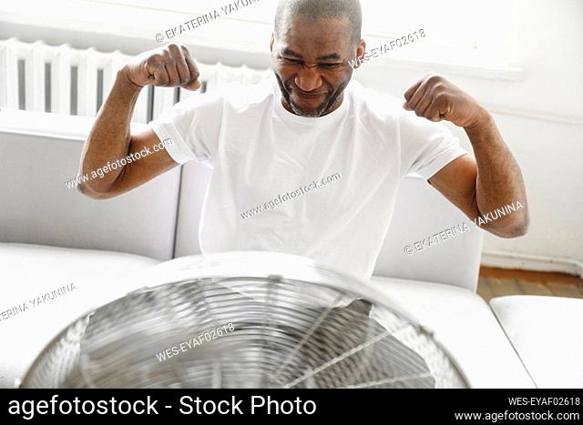 Man flexing muscles in front of electric fan at home