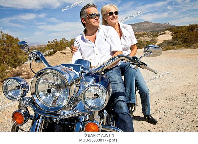 Senior couple wear sunglasses seated on motorcycle on desert road