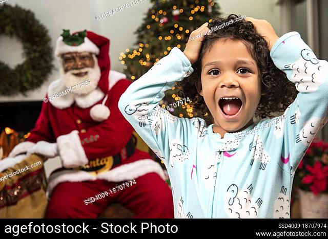 Cute girl in pajamas screaming in surprise as she catches Santa Claus delivering gifts. Black Santa
