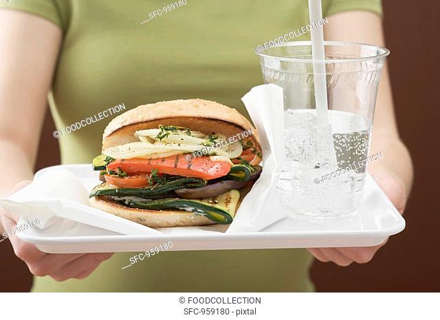 Woman holding roll filled with grilled vegetables & soda on tray