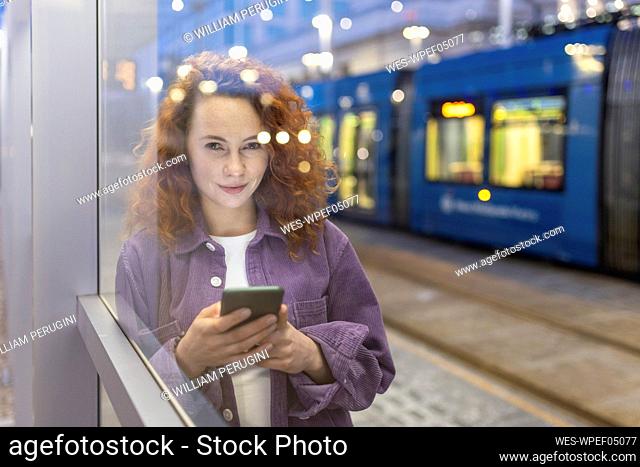Beautiful woman holding mobile phone looking through glass at tram station