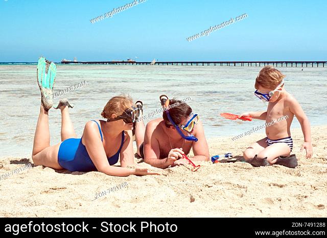 Mother, father and little son on summer vacation at the seaside, They wearing snorkels and flippers while lying and playing on the beach