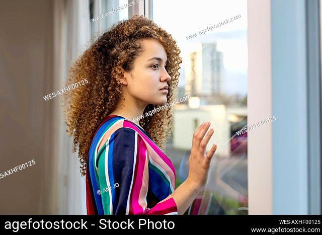 Young woman with curly hair looking through window at home
