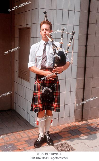 Man standing in street wearing traditional Scottish clothing playing bagpipes