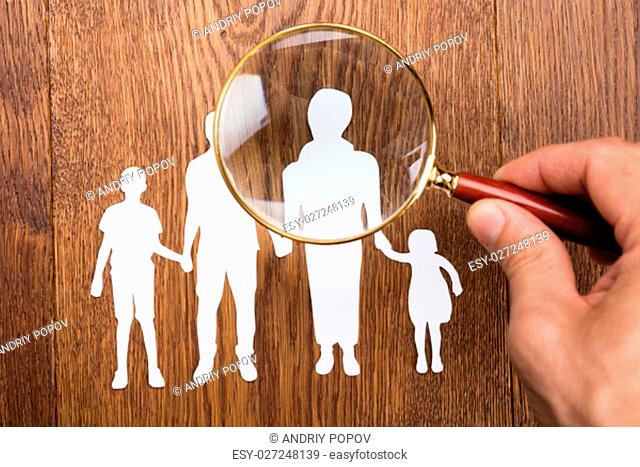 High Angle View Of Person Using Magnifying Glass On Family Papercut At Wooden Desk