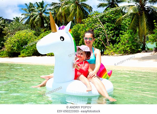 Smiling Portrait Of Mother And Daughter Enjoying Sitting On White Unicorn Float At Beach