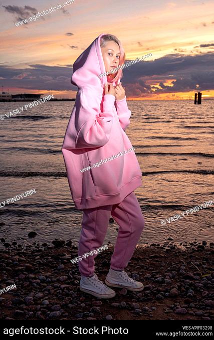 Portrait of young woman wearing pink hooded shirt standing on beach at sunset