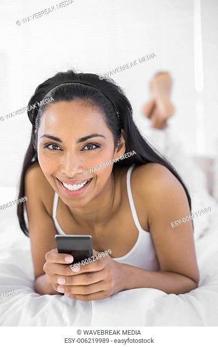 Beautiful smiling woman holding her smartphone lying on her bed